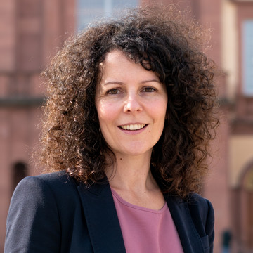 Angela Dörflinger hat dunkle, lockige Haare. Sie trägt einen schwarzen Blazer und ein fliederfarbenes Oberteil.