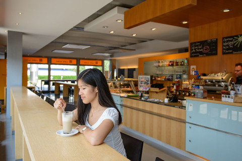 Eine Studentin trinkt einen Cappuccino im "Café Soleil" der Universität Mannheim und im Hintergrund steht ein Mitarbeiter hinter dem Thresen.