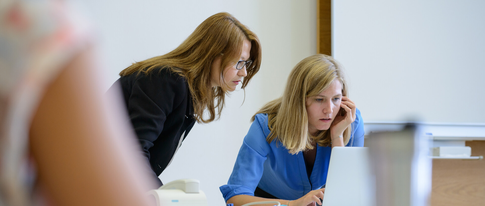 Zwei Frauen schauen zusammen auf den Display eines Laptops. Sie stehen vorne an der Tafel.