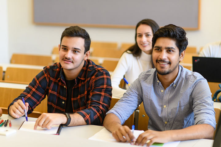 Zwei Studenten sitzen an einem Tisch in einem Seminarraum. Vor ihnen liegen Papier und Stifte.