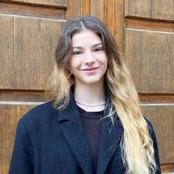 Helen Kalmbach steht vor einem runden hölzernen Tor der Uni Mannheim. Sie trägt einen schwarzen langen Wollmantel und hat offene lange Haare.