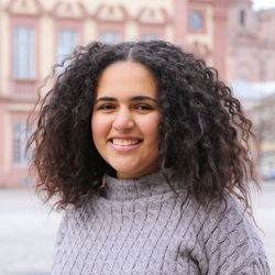 Miral Elsayed hat schwarzes schulterlanges lockiges Haar und trägt einen grauen Pulluvor mit Strickmuster. Sie lächelt und steht auf dem Ehrenhof.