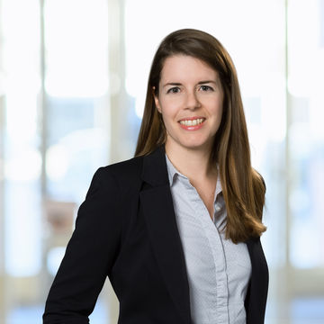 Linda Schädler hat lange braune Haare. Sie trägt einen schwarzen Blazer und ein graues Hemd mit weißen Punkten.