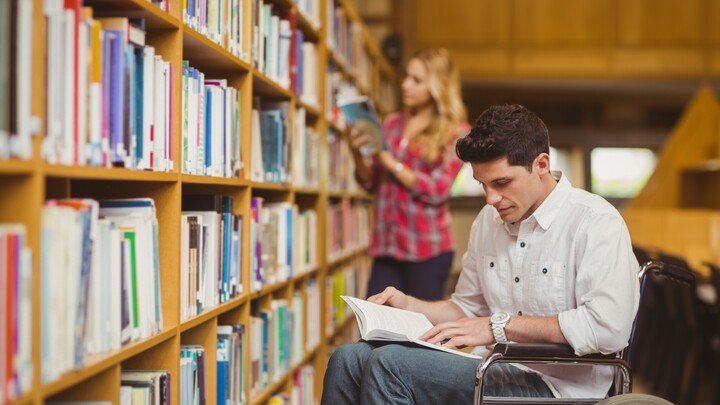 Student in wheelchair talking with classmate Ein Studierender im Rollstuhl mit einem Buch auf dem Schoß vor einem Bücherregal. Link: Beratung für behinderte und chronisch kranke Studierende